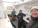 Three people bundled in cold-weather gear smiling and working inside a laboratory space.