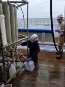 Two people in hard hats collect seawater samples from a CTD rosette on the deck of a research vessel.