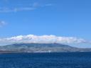 The coastal city of Sao Miguel at the base of a green mountain partly covered by clouds, viewed from across the ocean under a clear blue sky.