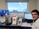 A researcher in a white lab coat works at a computer beside flow-cytometry equipment, with snowy Antarctic mountains and the red research vessel RV Laurence M. Gould visible through the window behind them.
